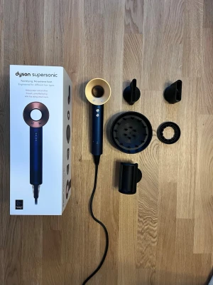 Dyson Supersonic hårfön blå/guld - Säljer en Dyson Supersonic hårfön i snygg blå och guld färg. Medföljer fyra olika munstycken för styling och volym, samt originalkartong. Hårfönen har en modern, rund design och är tillverkad i plast och metall. Perfekt för dig som vill ha ett proffsigt stylingverktyg hemma.
