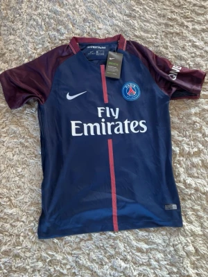 PSG Neymar Jr matchtröja Nike - Snygg PSG matchtröja i mörkblått med vinröda ärmar och röda detaljer. Neymar Jr och nummer 10 på ryggen. Tillverkad i lätt och ventilerande material, perfekt för fotboll eller träning. Fly Emirates-logga på bröstet och Nike-logga på framsidan.