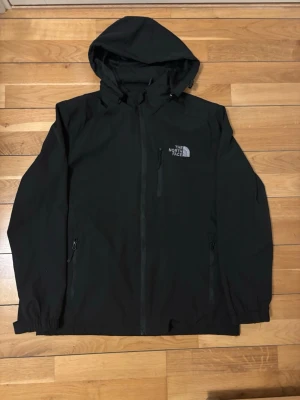 Svart vindjacka från The North Face - Snygg svart vindjacka från The North Face med huva och dragkedja framtill. Jackan har två sidofickor med dragkedja, justerbara muddar och logga på bröstet och ryggen. Perfekt för blåsiga dagar och enkel att matcha med allt.