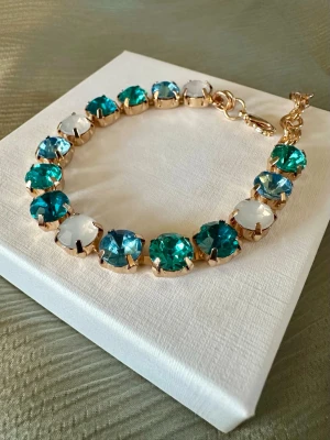 Armband med blå och vita stenar - Snyggt armband i blått, turkost och vitt, infattade i guldfärgad metall. Armbandet har justerbar kedja och ger en lyxig känsla med sina glittrande stenar. Perfekt accessoar för att lyfta din outfit. Justerbart ca 17-22 cm. Asken på bilden följer inte med (skickas i annan ask).