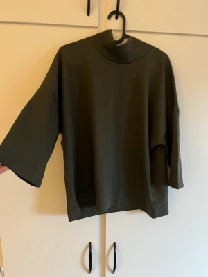 Olivgrön loose topp med trekvartsärm - Snygg olivgrön topp med hög hals och vida trekvartsärmar. Modellen är loose fit och har en minimalistisk design utan synliga detaljer. Perfekt för en chill och stilren look. Från Carin Wester. Katt finns i hemmet.