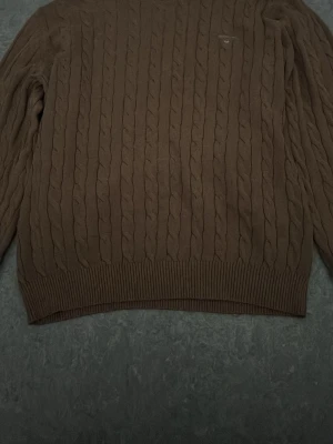 Brun kabelstickad tröja från GANT. PRIS KAN DISKUTERAS! - Snygg brun kabelstickad tröja från GANT i premium bomull. Tröjan har rund halsringning, ribbade muddar och diskret GANT-logo på bröstet. Perfekt för dig som gillar klassisk och stilren look.