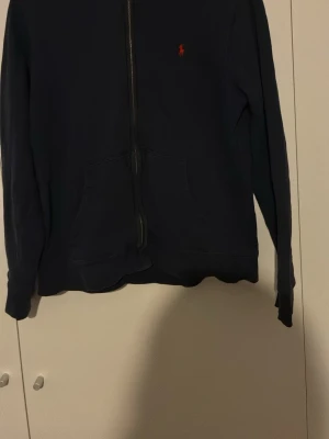 Mörkblå hoodie från Polo Ralph Lauren - Mörkblå hoodie med dragkedja från Polo Ralph Lauren. Klassisk design med röd broderad logga på bröstet och känguruficka framtill. Tillverkad i mjukt bomullsmaterial som är skönt mot huden. Perfekt för en avslappnad och stilren look.