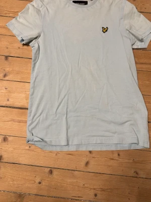 Ljusblå t-shirt från Lyle & Scott - Snygg ljusblå t-shirt från Lyle & Scott med klassisk gul logga på bröstet. T-shirten har rund halsringning och korta ärmar. Tillverkad i mjuk bomull för en skön känsla. Perfekt till jeans eller shorts för en clean look.