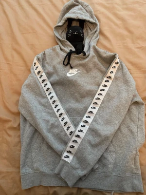 Grå Nike hoodie med logoband - Snygg grå hoodie från Nike med vit logga på bröstet och vita band med svarta Nike-loggor längs ärmarna och axlarna. Klassisk känguruficka framtill och justerbar huva med snören. Perfekt för en sportig och avslappnad stil.