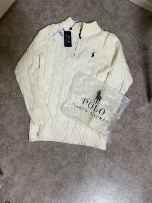 Vit stickad tröja från Polo Ralph Lauren - Vit kabelstickad tröja från Polo Ralph Lauren med halv dragkedja och broderad logga på bröstet. Tröjan har ribbade muddar och hög krage, perfekt för lager-på-lager. Klassisk och stilren design som funkar till det mesta.