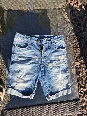 Blå slitna jeansshorts Adrian Hammond - Säljer ett par blå jeansshorts från Adrian Hammond med slitna detaljer och upprullade benslut. Shortsen har klassisk femficksdesign, knappar med logga och en cool, avslappnad look. Perfekta för sommaren och ger en trendig streetstyle-vibe.
