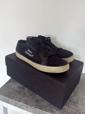 Svarta Saint Laurent sneakers - Svarta sneakers från Saint Laurent med vit logga på sidan. Skorna ska ha slitningar orginal. Men dom har används en del och därför är det mer slitningar på dom än vad det ska vara. Trendiga och snygga skor som går att använda till nästan allt. Nypris runt 7000, mitt pris 1500