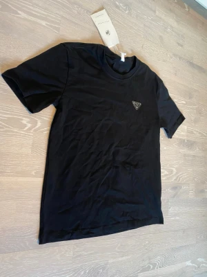 Svart t-shirt från Prada - Snygg svart t-shirt från Prada med klassisk rund hals och korta ärmar. På bröstet sitter Pradas ikoniska trekantslogga i metall och läder. Perfekt för dig som gillar stilrena och exklusiva plagg.