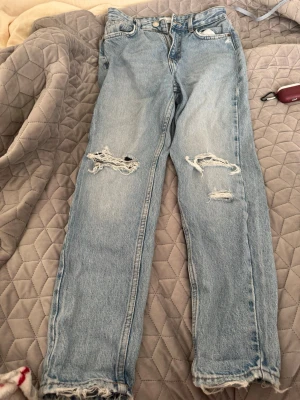 Ljusblå slitna mom jeans - Säljer ett par ljusblå mom jeans med slitningar och hål på båda benen. Jeansen har hög midja, raka ben och råa kanter vid bensluten. Perfekta för en avslappnad och trendig look. Klassisk femficksmodell i mjuk denim.