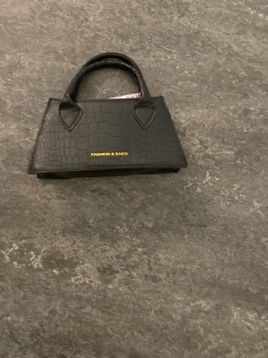 Svart handväska med krokoprägling - Snygg svart handväska från Fashion & Bags med trendig krokopräglad yta. Väskan har två handtag och en stilren, avsmalnande form. Perfekt accessoar för att lyfta din outfit med en lyxig känsla.