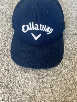 Mörkblå Callaway keps - Snygg mörkblå keps från Callaway med broderad logga framtill och Odyssey-detalj baktill. Justerbar rem baktill för perfekt passform. Perfekt accessoar för golfbanan eller en sportig look.