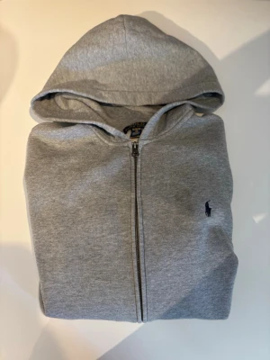 Grå hoodie från Polo Ralph Lauren XL kids size (170cm) - Snygg grå hoodie från Polo Ralph Lauren med dragkedja framtill och klassisk huva. Tröjan har en liten broderad logga på bröstet och är tillverkad i mjukt bomullsmaterial. Perfekt för en avslappnad och stilren look. XL kids size 