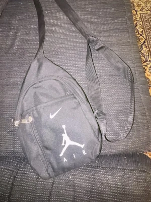 Svart Jordan Air ryggsäck - Säljer en svart ryggsäck från Nike Jordan med klassisk Air-logga och vit swoosh på framsidan. Väskan har justerbar axelrem, dragkedjefack fram och bak samt är tillverkad i slitstark textil. Perfekt för dig som gillar sportig och stilren design.