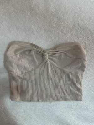 Beige ribbad bandeau tubtopp - Säljer en beige ribbad bandeau tubtopp med twistad detalj framtill. Toppen är axelbandslös och har en mjuk, stretchig passform som sitter snyggt på kroppen. Samt finns de gummi som gör att tröjan inte åker ner och visar inte för mycket. Perfekt att styla till jeans eller kjol för en trendig look.