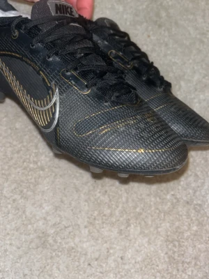 Nike Mercurial 14 elite AG - Säljer ett par Nike Mercurial fotbollsskor i svart med snygga gulddetaljer och silverfärgad swoosh. Skorna har snörning, låg profil och mönstrad ovandel i syntetmaterial för bra bollkontroll. Perfekta för dig som vill sticka ut på planen.