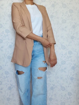 Beige oversized kavaj med 3/4-ärm - Stilren beige kavaj i oversized modell med 3/4-ärm och klassisk krage. Kavajen har två fickor framtill och är tillverkad i ett mjukt, lätt material som ger en avslappnad men trendig look. Perfekt att slänga över en t-shirt för en cool outfit.