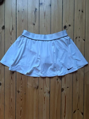Vit sportig skort från Nike - Supersnygg vit skort från Nike med bred resår i midjan och inbyggda shorts under kjolen. Tillverkad i ett lätt och stretchigt material som andas, perfekt för träning eller sportiga outfits. Diskret Nike-logga broderad framtill.