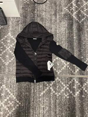 Svart Moncler cardigan  - Snygg svart Moncler cardigan  från Moncler med quiltad front och huva i glansigt material. Ärmarna är i mjuk stickad bomull och ena ärmen har en ficka med dragkedja samt Moncler-logga. Perfekt för dig som vill ha en exklusiv streetwear-look.