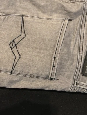Grå slim fit jeans från Replay - Säljer ett par gråa slim fit jeans från Replay, modell Anbass. Jeansen har en unik svart broderad detalj på bakfickan och klassisk femficksdesign. Perfekta för dig som gillar stilrena men ändå personliga jeans.