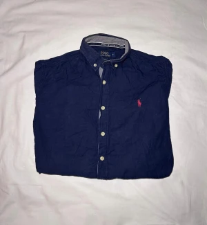 Ralph Lauren | Skjorta | M - Klassisk marinblå skjorta från Polo Ralph Lauren med knappar framtill och ikoniska röda logobroderiet på bröstet. Skjortan har lång ärm, button-down krage och är gjord i mjuk bomull. Perfekt till jeans eller chinos för en clean och stilren look. Mått: 65 Längd, 56 Bredd
