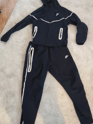 Mörkblå Nike loungewear-set - Mörkblått loungewear-set från Nike med vit piping och dragkedja på tröjan. Setet består av en hoodie med fickor och matchande byxor med dragsko och vita detaljer längs benen. Perfekt för chill dagar hemma eller när du vill vara bekväm och snygg. Äkta såklart köpt för 2900
