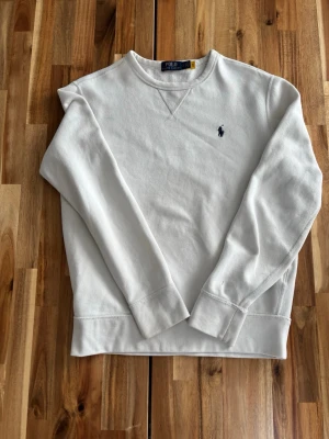 Vit sweatshirt från Polo Ralph Lauren - Klassisk vit sweatshirt från Polo Ralph Lauren med rund hals och diskret broderad logga på bröstet. Tröjan har ribbade muddar vid ärmslut och nederkant, samt är tillverkad i mjukt bomullsmaterial. Perfekt för en avslappnad och stilren look. Det finns en blå fläck uppe till vänster, liten svårt att se.