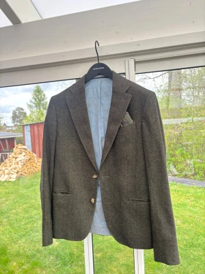 Mörkgrå ullkavaj från sand - Stilren mörkgrå kavaj från sand i ullmaterial. Kavajen har två knappar framtill, klassiska slag och ljusblått mönstrat innerfoder. Perfekt för en dressad look och snygg att matcha med skjorta eller t-shirt.