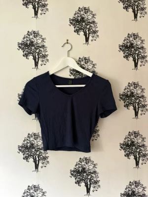 Mörkblå croppad ribbad topp - En mörkblå croppad topp med korta ärmar och v-ringning. Toppen är ribbad och har en slim passform som framhäver figuren. Perfekt att matcha med höga jeans eller kjol.