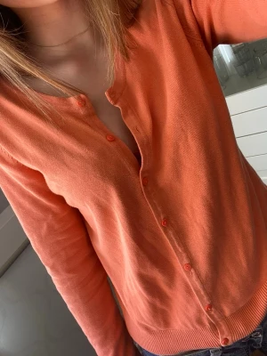 Korall/peach-färgad kofta - Snygg orange kofta med rund halsringning och matchande knappar framtill. Långärmad och i mjukt, tunt stickat material som passar perfekt till lager-på-lager. Enkel och stilren design som är lätt att matcha.