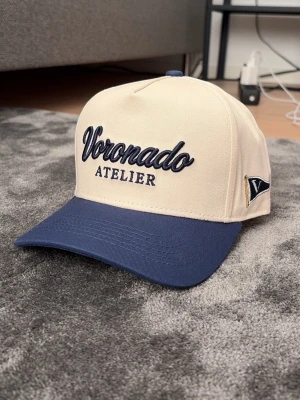 Voronado atelier keps  - Snygg keps från Voronado Atelier i beige med marinblå skärm och broderad logga framtill. Klassisk sexpanelsdesign med böjd skärm och justerbar passform. Perfekt accessoar för en stilren och avslappnad look.