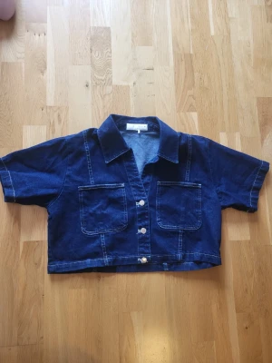 Mörkblå croppad jeansjacka/topp i denim - Snygg croppad jeansjacka/topp i mörkblå denim med korta ärmar, två stora bröstfickor och silvriga knappar framtill. Klassisk skjortkrage och markerade sömmar ger en cool look. Perfekt att styla med höga jeans eller kjol.