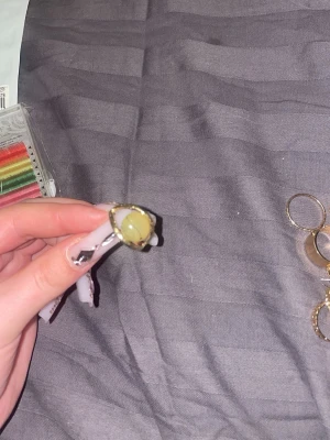 Guldfärgad ring med grön pärla - Snygg guldfärgad ring med en stor rund grön pärla i mitten. Ringen har en enkel men iögonfallande design och passar perfekt för dig som vill sticka ut. Materialet ser ut att vara metall med en glansig yta och en pärlliknande sten.