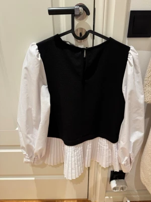 Svartvit blus med plisserad nederdel - Trendig svartvit blus med långärmade vita puffärmar och plisserad vit nederdel. Överdelen är svart och har en rak, boxig passform. Perfekt för dig som gillar kontraster och stilrena detaljer.
