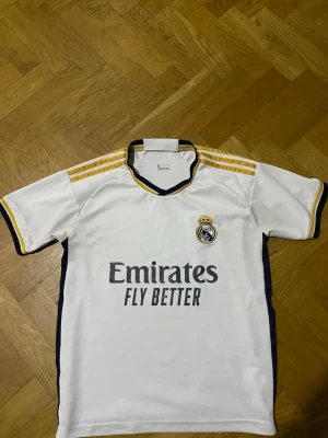 Real Madrid matchtröja vit/gul - Snygg vit Real Madrid matchtröja från Adidas med gula detaljer och klubbens broderade logga på bröstet. Tröjan har svarta och gula ränder på axlarna, korta ärmar och trycket 'Emirates Fly Better' på framsidan. Perfekt för fotbollsträning eller att bära på läktaren.