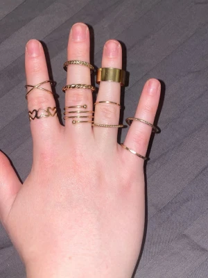 Set med guldiga ringar i olika former - Säljer ett set med flera tunna guldiga ringar i olika former och mönster. Några ringar har tvinnad design, hjärtan, korsade band och en bredare slät ring. Perfekt för att mixa och matcha på olika fingrar.