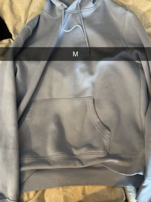 Ljusblå hoodie med magficka - En stilren ljusblå hoodie med klassisk magficka och justerbar huva med dragsko. Hoodien har en relaxed fit och är tillverkad i mjukt bomullsmaterial. Perfekt för en chill och avslappnad look.