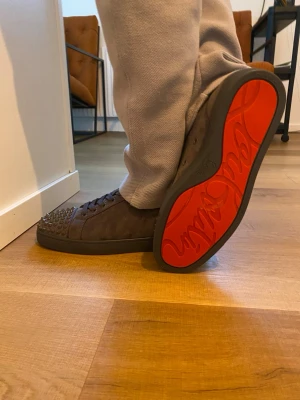 Gråa Christian Louboutin sneakers - Snygga gråa sneakers från Christian Louboutin i mocka med svarta nitar på tån. Skorna har klassisk låg modell, svarta snören och svart sula. Perfekta för dig som vill sticka ut med exklusiva detaljer och lyxig känsla.