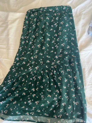 Grön blommig långkjol med volang - Supersöt långkjol i mörkgrönt tyg med små rosa och gula blommor. Kjolen har volangdetalj och är perfekt för vår och sommar. Materialet känns mjukt och lätt, troligen bomull eller plolyster. Pris kan diskuteras🥰