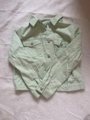 Ljusgrön jeansjacka från HM - Snygg ljusgrön jeansjacka från HM med klassisk krage, två bröstfickor med knappar och raka ärmar. Jackan är tillverkad i ett mjukt denimtyg och har en avslappnad passform. Perfekt för att lyfta din outfit med en fräsch färg. Den är i storlek L men upplevs som S