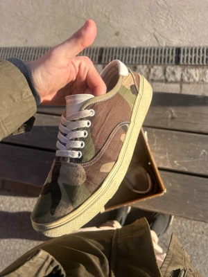 Saint Laurent camo sneakers - 🔥 Saint Laurent Paris sneakers i camouflage Storlek: 43 Made in Spain 🇪🇸  Säljer ett par riktigt snygga Saint Laurent low-top sneakers i klassiskt camo-mönster. Perfekta om du vill ha en clean men exklusiv sneaker som sticker ut lagom mycket.  • Original Saint Laurent Paris • Canvas med läderdetaljer • Bekväma och lätta att matcha • Bra skick med naturliga tecken på användning • Cream/vita snören och sula • Storlek 43  Passar perfekt till både streetwear och mer clean outfits. 