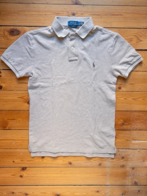 Beige pikétröja från Polo Ralph Lauren - Klassisk beige pikétröja från Polo Ralph Lauren med korta ärmar, krage och knappar framtill. Tröjan har den ikoniska broderade loggan på bröstet och är tillverkad i bomull för en bekväm känsla.