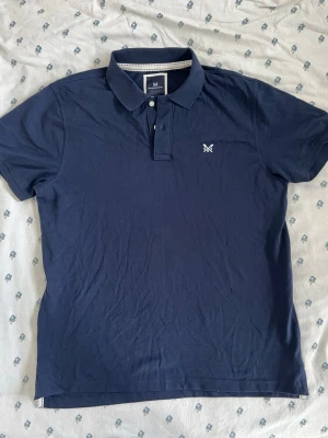 Marinblå pikétröja från Crew Clothing Company - Säljer en klassisk marinblå pikétröja från crew clothing company storlek XL med korta ärmar och krage. Tröjan har två knappar framtill och den ikoniska Morris-loggan broderad på bröstet. Tillverkad i mjuk bomull som känns skön mot huden.