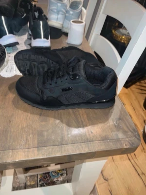 Svarta sneakers från Fila - Snygga helsvarta sneakers från Asics med klassisk låg profil. Skorna har ovandel i mesh och mocka, samt svarta detaljer och snörning. Perfekta för en stilren och sportig look.