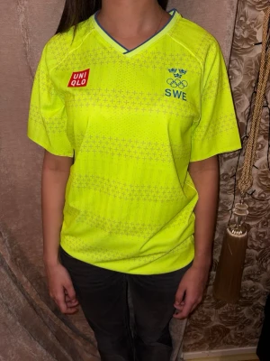 Gul Uniqlo SWE sporttröja - Sportig gul t-shirt från Uniqlo med blå detaljer och SWE-logga på bröstet. Tröjan har v-ringning, korta ärmar och ett diskret mönster över hela tyget. Perfekt för dig som gillar sportig och färgstark stil. Priset går att diskuteras 