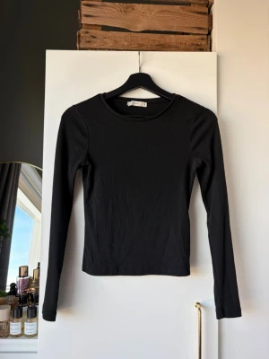 Svart långärmad topp från Pigalle - En svart, ribbad långärmad topp från Pigalle i storlek XS. Toppen har en rund halsringning och är figurnära i modellen, perfekt att styla med jeans eller kjol. Enkel och stilren design som funkar till många olika looks.