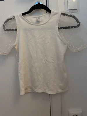 Vit t-shirt med puffärmar i mesh - Supersöt vit t-shirt med korta puffärmar i transparent mesh och små prickar. Klassisk rund halsringning och rak passform. Perfekt för dig som vill ha en t-shirt med det lilla extra och en trendig touch.