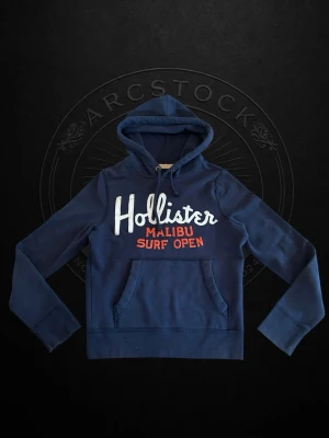 Hollister hoodie - Mörkblå hoodie från Hollister med vit och röd text på bröstet där det står 'Hollister Malibu Surf Open'. Klassisk känguruficka framtill och dragsko i huvan. Perfekt för en avslappnad och sportig stil.