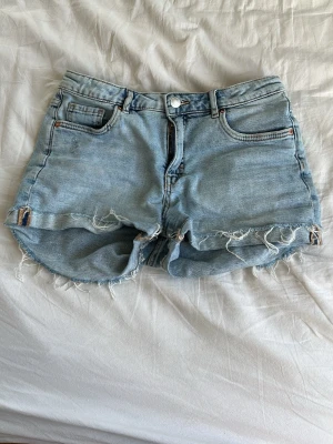 Ljusblå jeansshorts med fransig kant - Säljer ett par ljusblå jeansshorts med fransig, rå kant och klassisk femficksdesign. Shortsen har normal passform och är perfekta för varma dagar. Stängs med knapp och dragkedja framtill. Snygga att matcha med en enkel t-shirt eller crop top.
