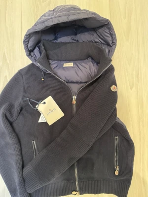 Mörkgrå pufferjacka från Moncler - Snygg mörkgrå pufferjacka från Moncler med stickade ärmar och kropp, samt vadderad huva och insida. Jackan har två dragkedjefickor, hög krage och klassisk Moncler-logga på ärmen. Perfekt för kalla vinterdagar och riktigt trendig look.